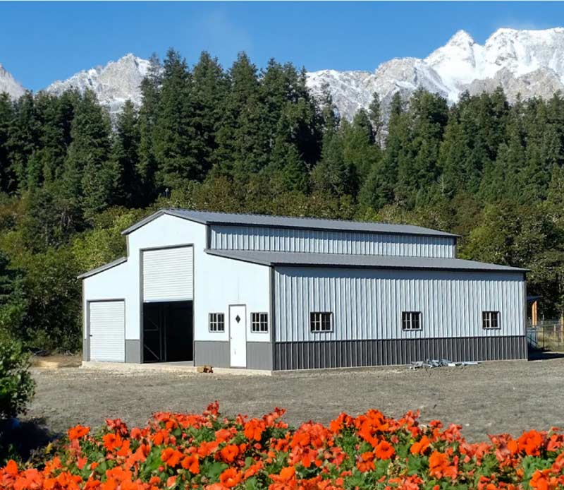 Large-scale grey and charcoal two-tone agricultural buildings with multiple roll-up doors and windows, situated in a rural landscape against a backdrop of evergreen trees and snow-capped mountains.