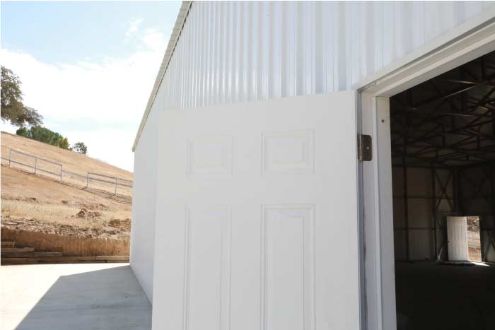 Close-up view of a white pedestrian door at the entrance of a newly constructed, high-ceiling, light-colored metal building or commercial carport