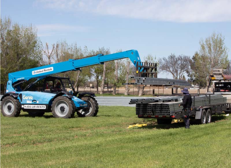 Heavy machinery (telehandler) lifting pre-fabricated garage building framing from a trailer at a residential or commercial job site on a grassy field
