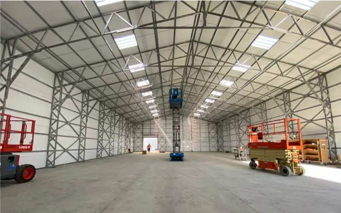 Interior view of a large, high-ceiling commercial metal building under construction, showing steel framing, roof trusses, skylights, concrete floor, and lift equipment.