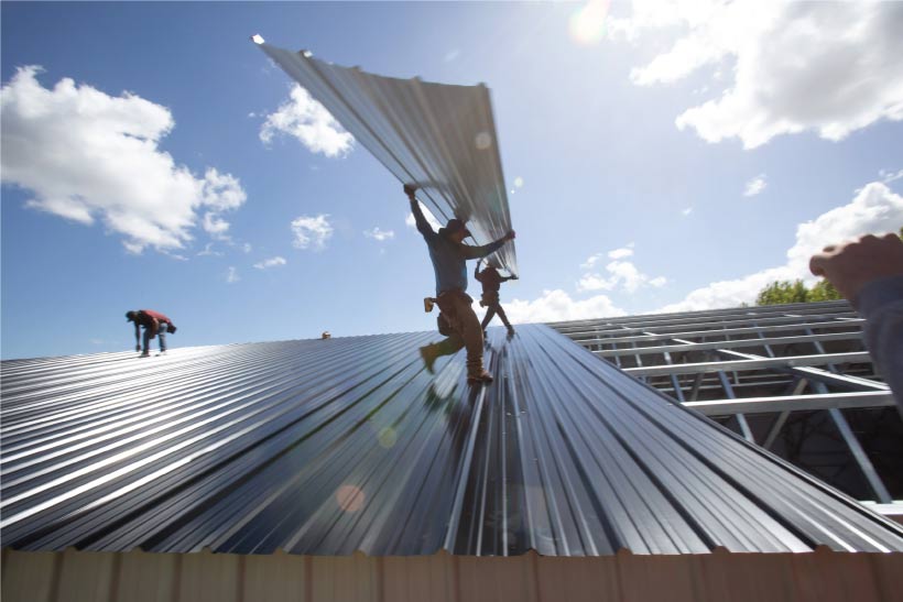 rew installing dark corrugated metal sheets on the roof of a large new metal building under a sunny, blue sky