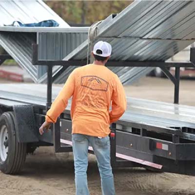 Quality Metal Carports worker in an orange long-sleeve shirt unloading manufactured metal sheets from a flatbed trailer at a construction site