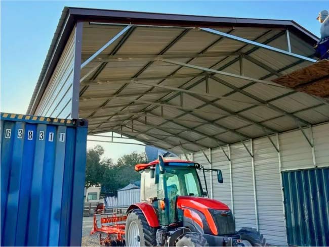 Agricultural metal canopy roof structure mounted between shipping containers providing shelter for a red farm tractor and equipment.