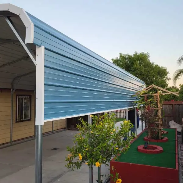 Blue metal carport with white framing and partial side panels installed over a concrete patio next to a garden planter with young trees.