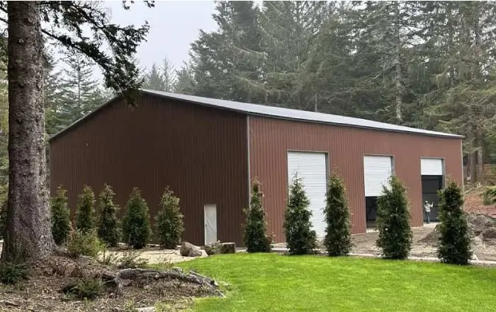 Large enclosed metal workshop building with three white roll-up doors and two-tone brown siding situated in a dense pine forest setting.