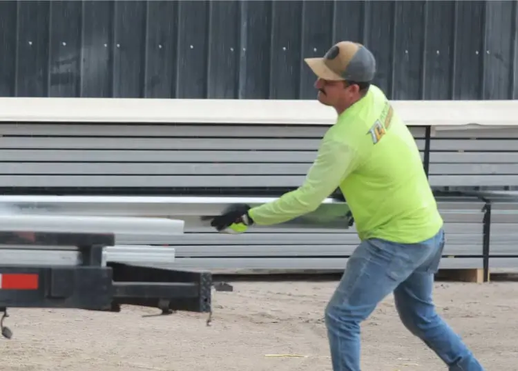 Construction worker in high-visibility gear handling long metal manufacturing components with stacks of steel sheet materials in the background.