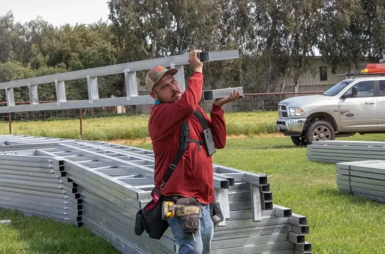 Installer carrying galvanized steel framing tubes on his shoulder at a construction site with stacks of metal trusses and a work truck in the background.