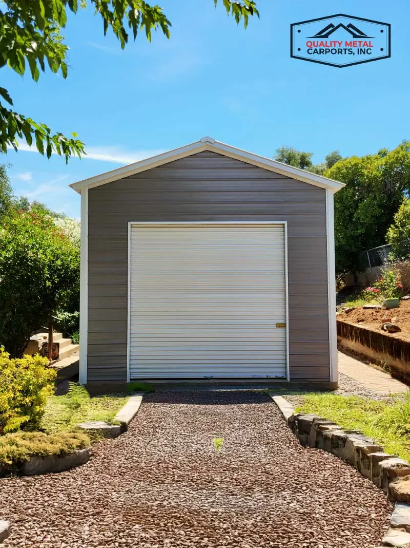Front view of a single-car prefab metal garage featuring grey horizontal siding, white trim, and a white roll-up door with a gravel driveway approach.