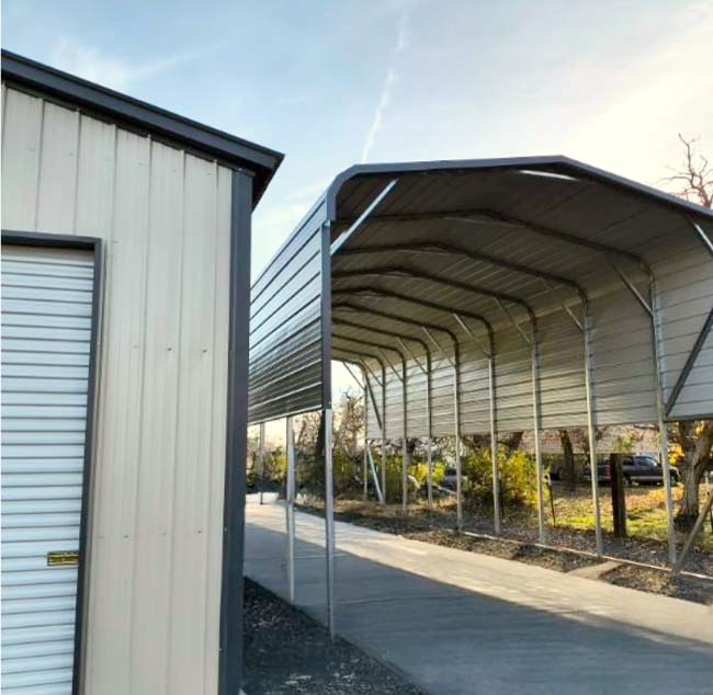 Heavy-duty open metal carport with a vertical roof design and reinforced steel trusses standing next to a fully enclosed beige metal garage building.