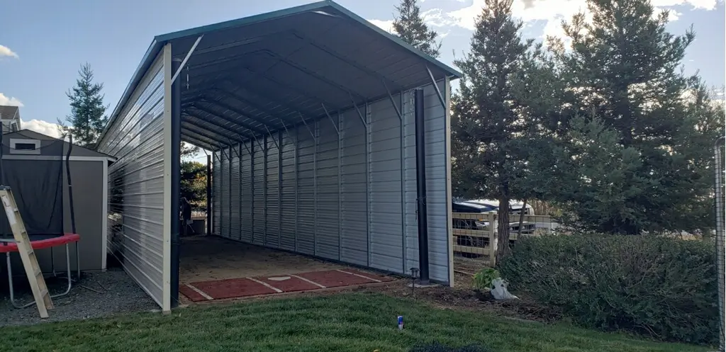 Tall grey metal RV carport with partial side paneling and high clearance installed in a residential backyard near a trampoline.